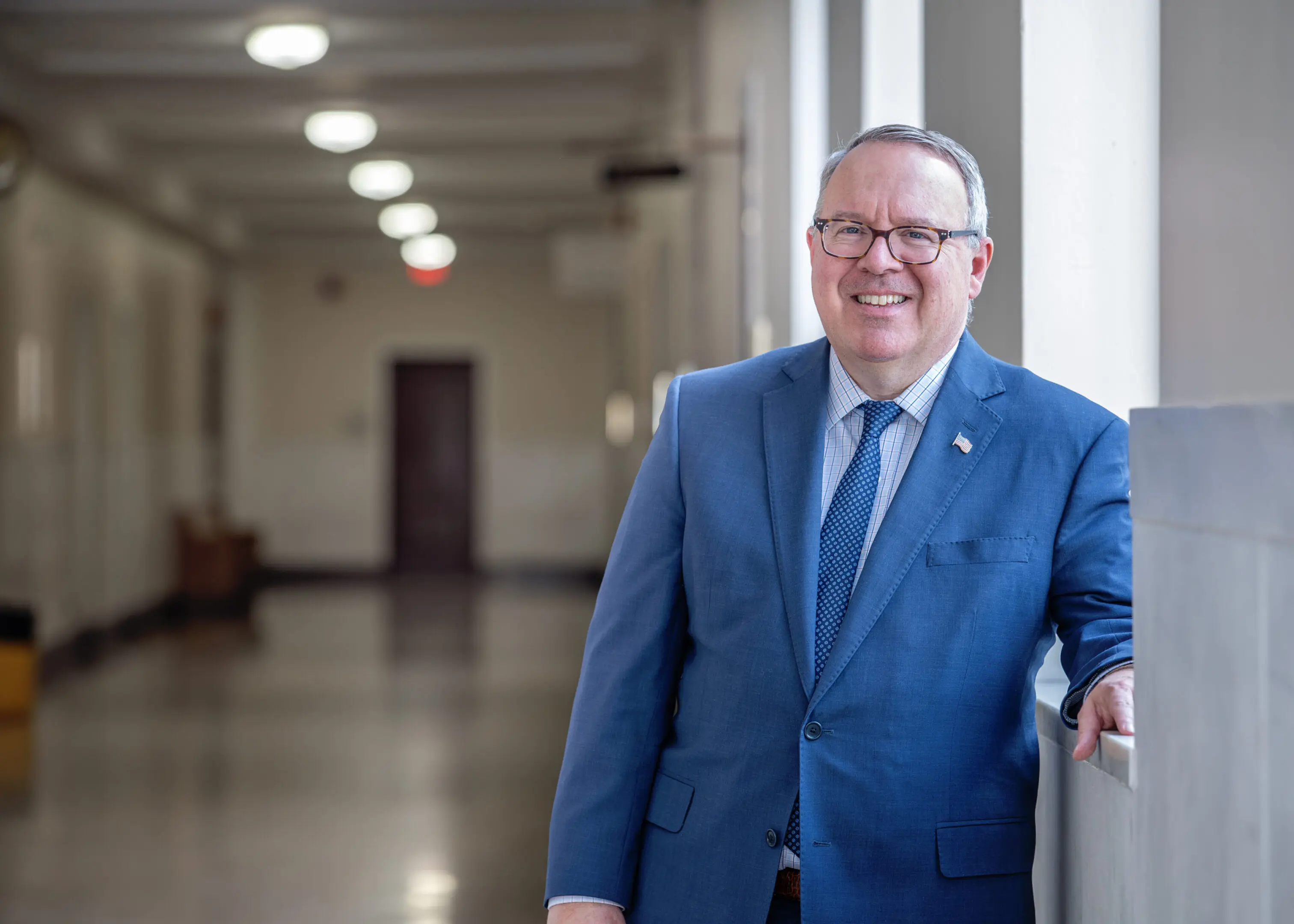 Smiling man in a blue suit standing in a hallway.