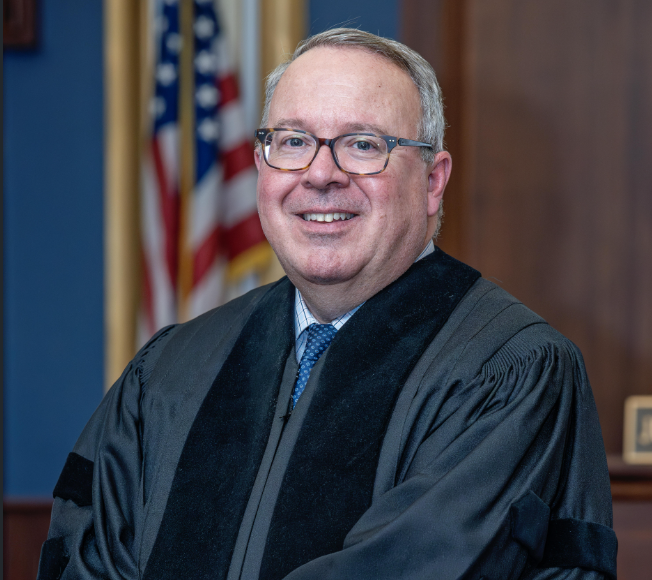 A smiling judge in a black robe with American flags in the background.