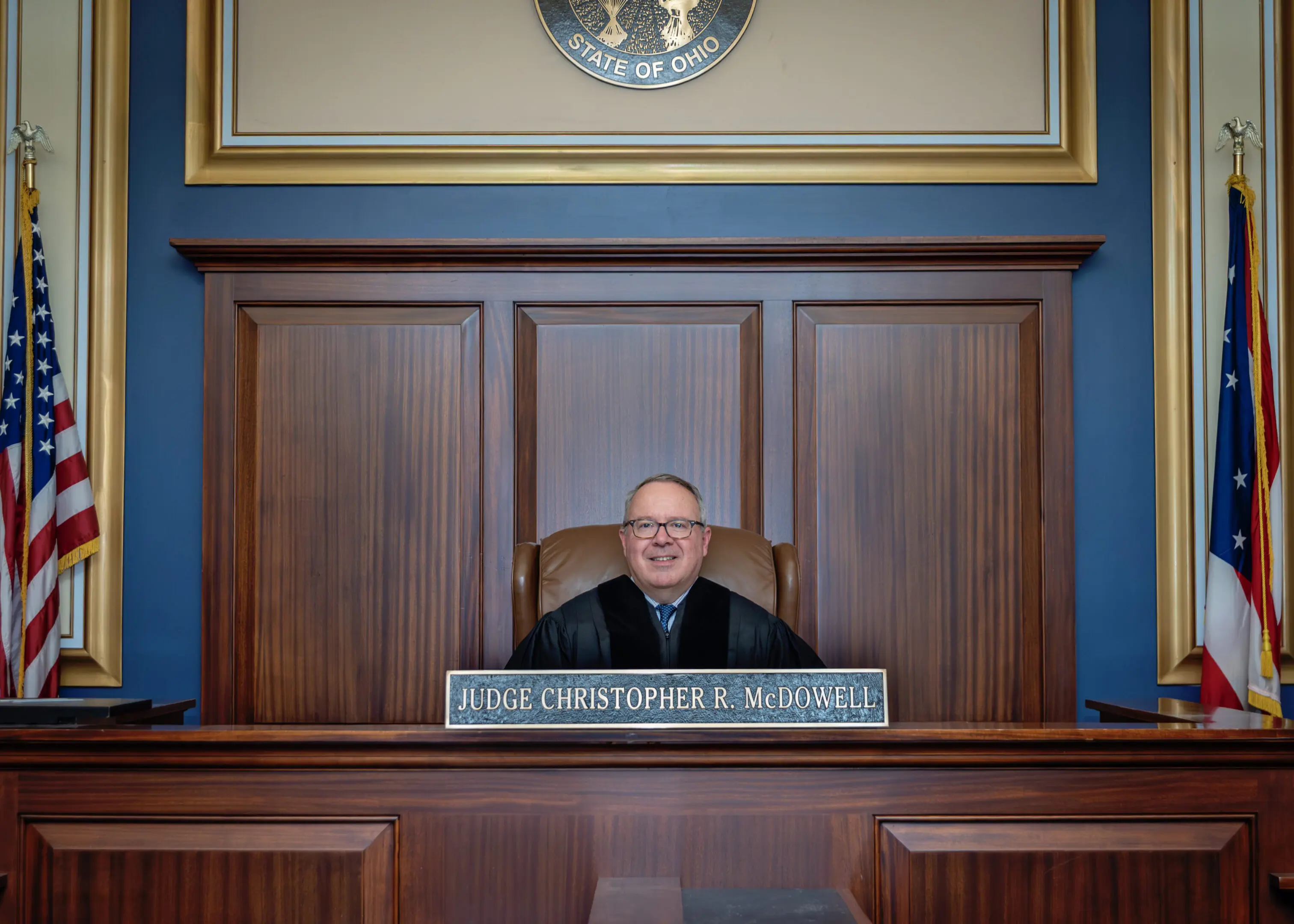 A smiling judge seated behind a wooden bench in a courtroom.